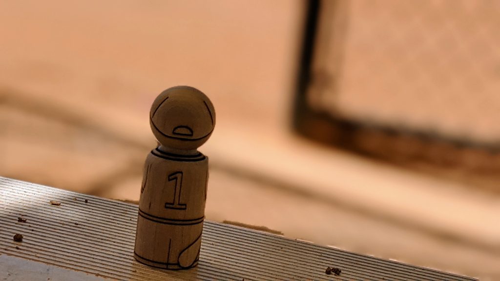 Wooden peg doll baseball player sitting in a baseball dugout on the bench looking out towards the dirt field.