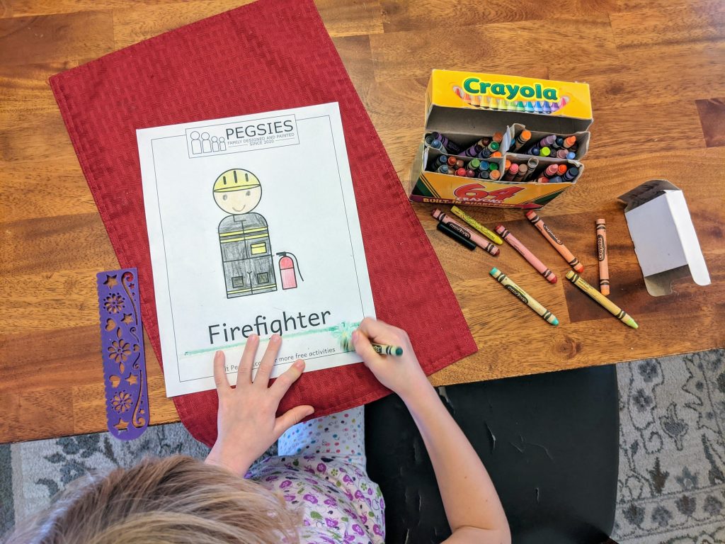 Little girl using crayons on a coloring page for a peg doll firefighter.